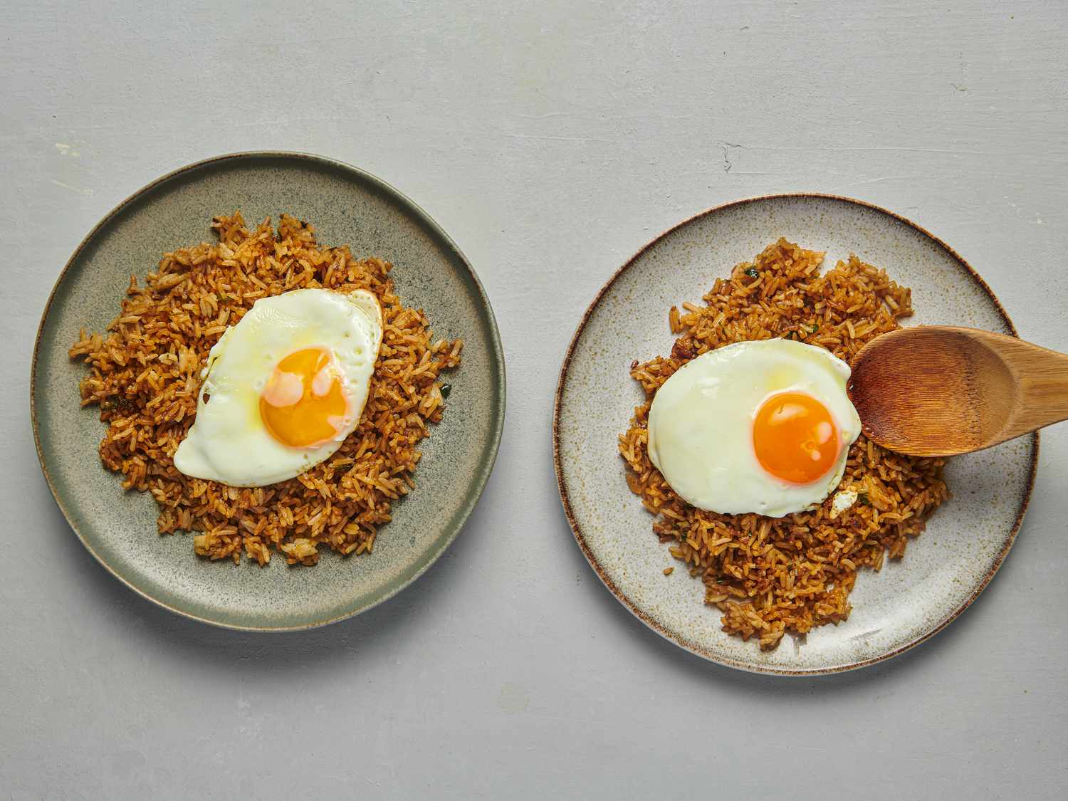 Two plates side by side, each containing a serving of fried rice which is topped with a sunny-side up fried egg. There is a wooden spatula over the right plate, depositing the egg on the rice.
