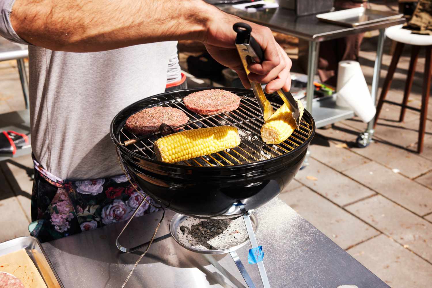 A person cooks corn and burgers on a small portable charcoal grill.