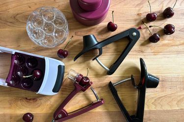Five cherry pitters on a wooden surface with a few cherries strewn around them