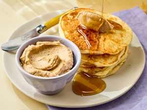 A plate of pancakes served with a small bowl of spreadable butter and honey with a yellowhandled knife nearby