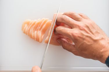 Two pairs of hands, one holding a knife and the other securing the chicken breast on a cutting board, with a chicken breast being thinly sliced.