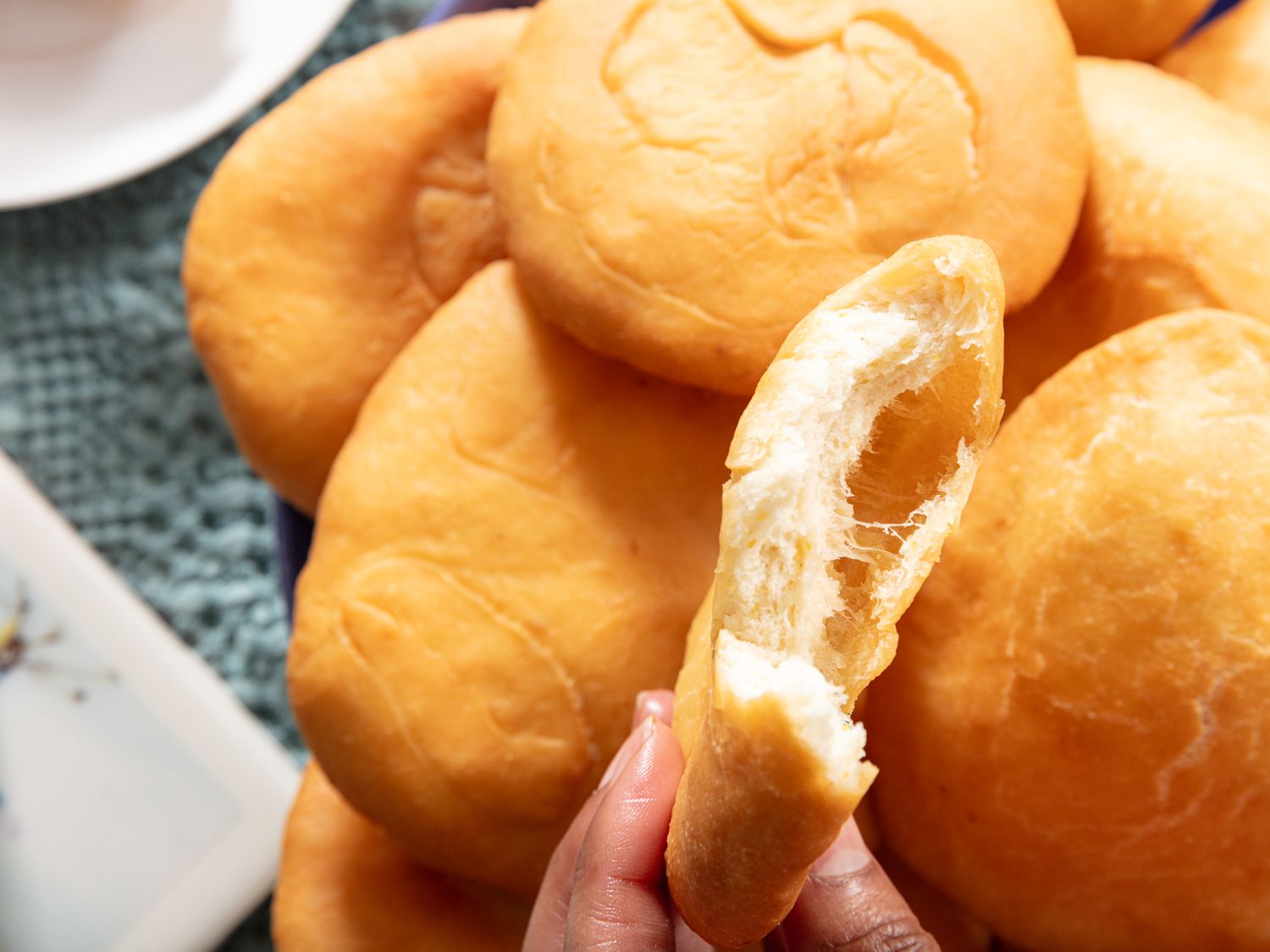 Close up of pile of johnny cakes one a platter, with a hand holding one bitten into in front. Platter is on a blue fabric surface 