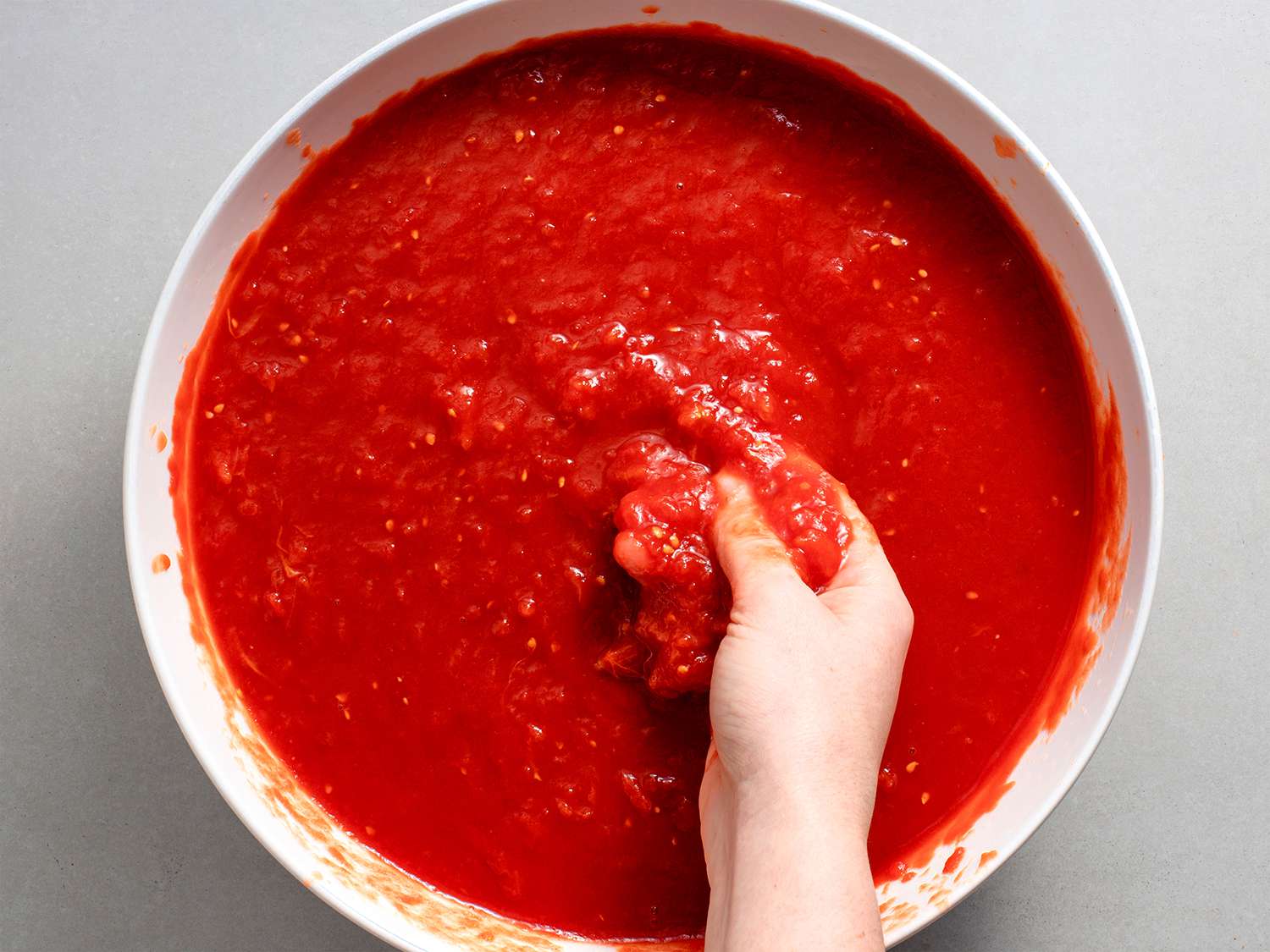 Tomatoes being crushed by hand in a white bowl.