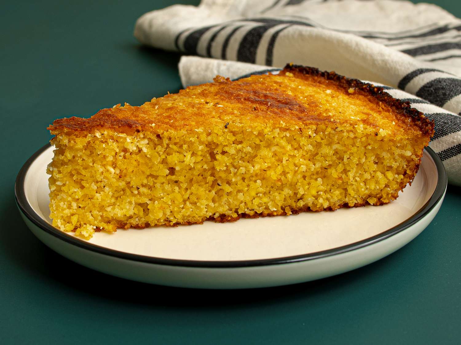 A slice of Southern-style unsweetened cornbread on a round white ceramic plate with a dark green rim. The cornbread is resting on a dark teal surface and there's a dishcloth in the background.