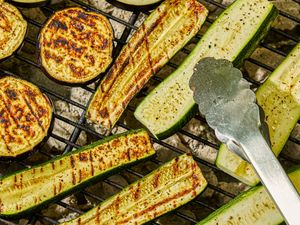 Slices of zucchini and eggplant grilling on a barbecue with metal tongs