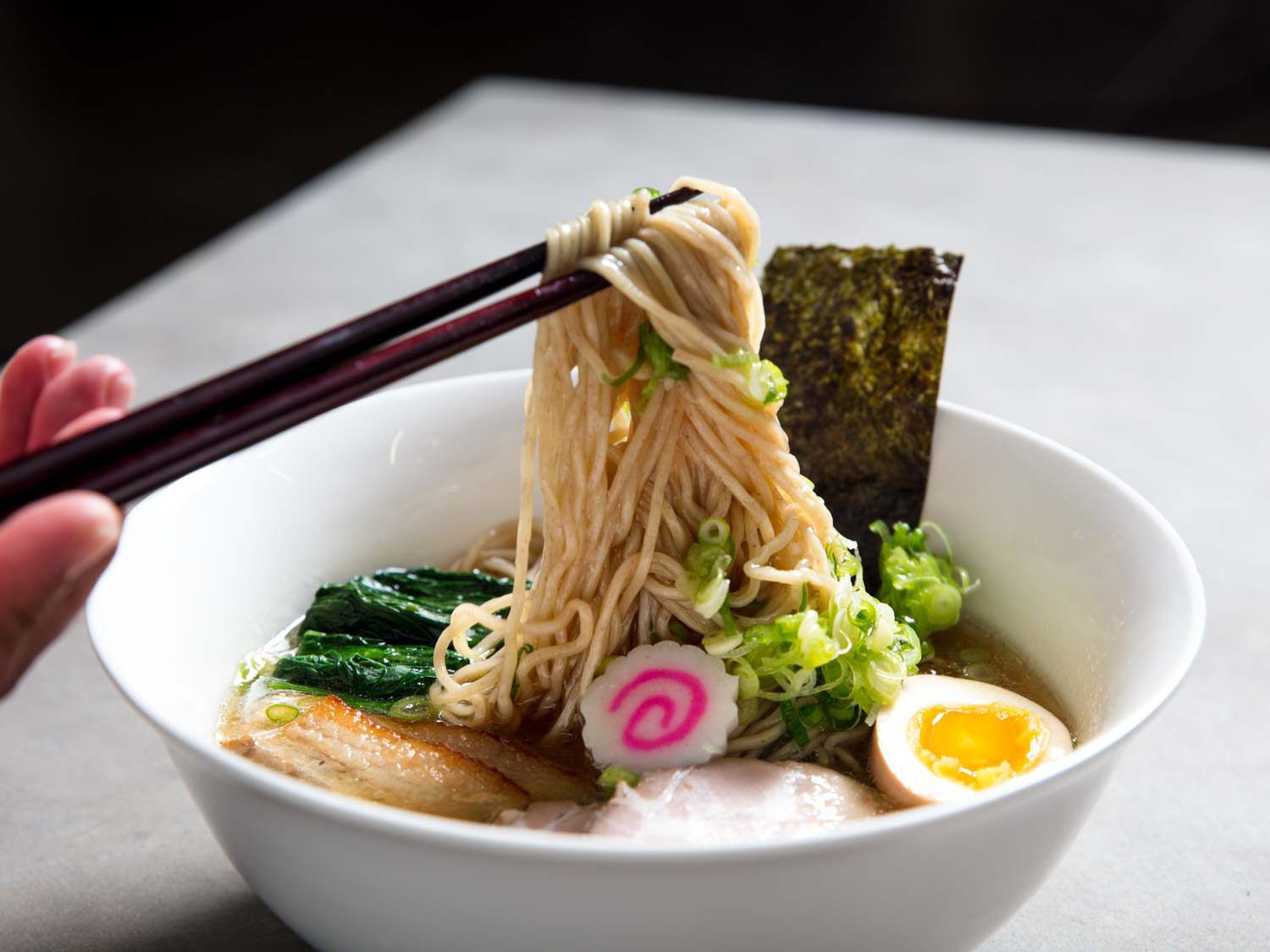 Chopsticks lifting a tangle of noodles from a bowl of shoyu ramen broth, seaweed, boiled egg, fish cake, scallions, and pork.