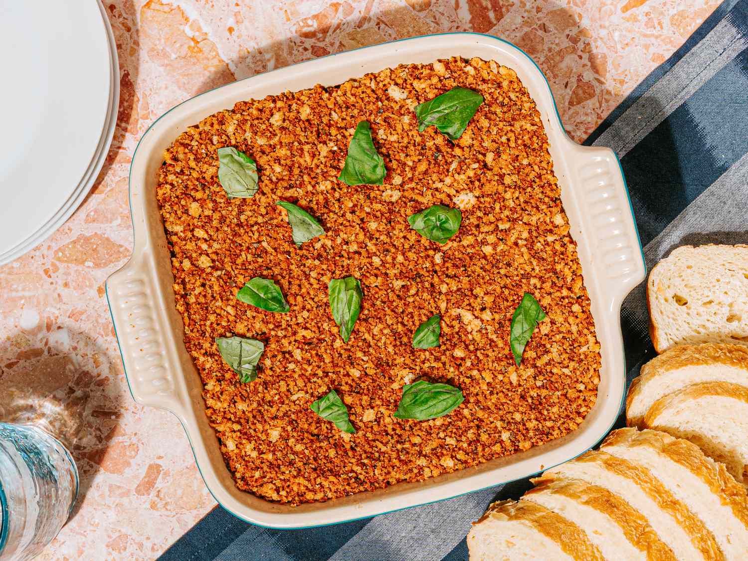 Baking dish containing baked dip with green herb leaves on top served with sliced bread on a striped tablecloth