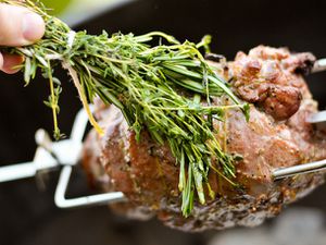 Boneless leg of lamb on a rotisserie, being basted with a bundle of fresh herbs. 