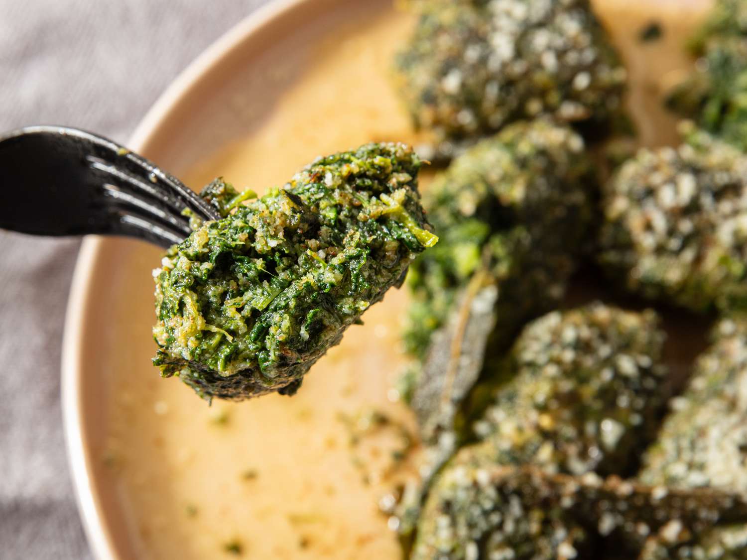 A fork holding a piece of cooked spinach dumpling with more dumplings on a plate in the background
