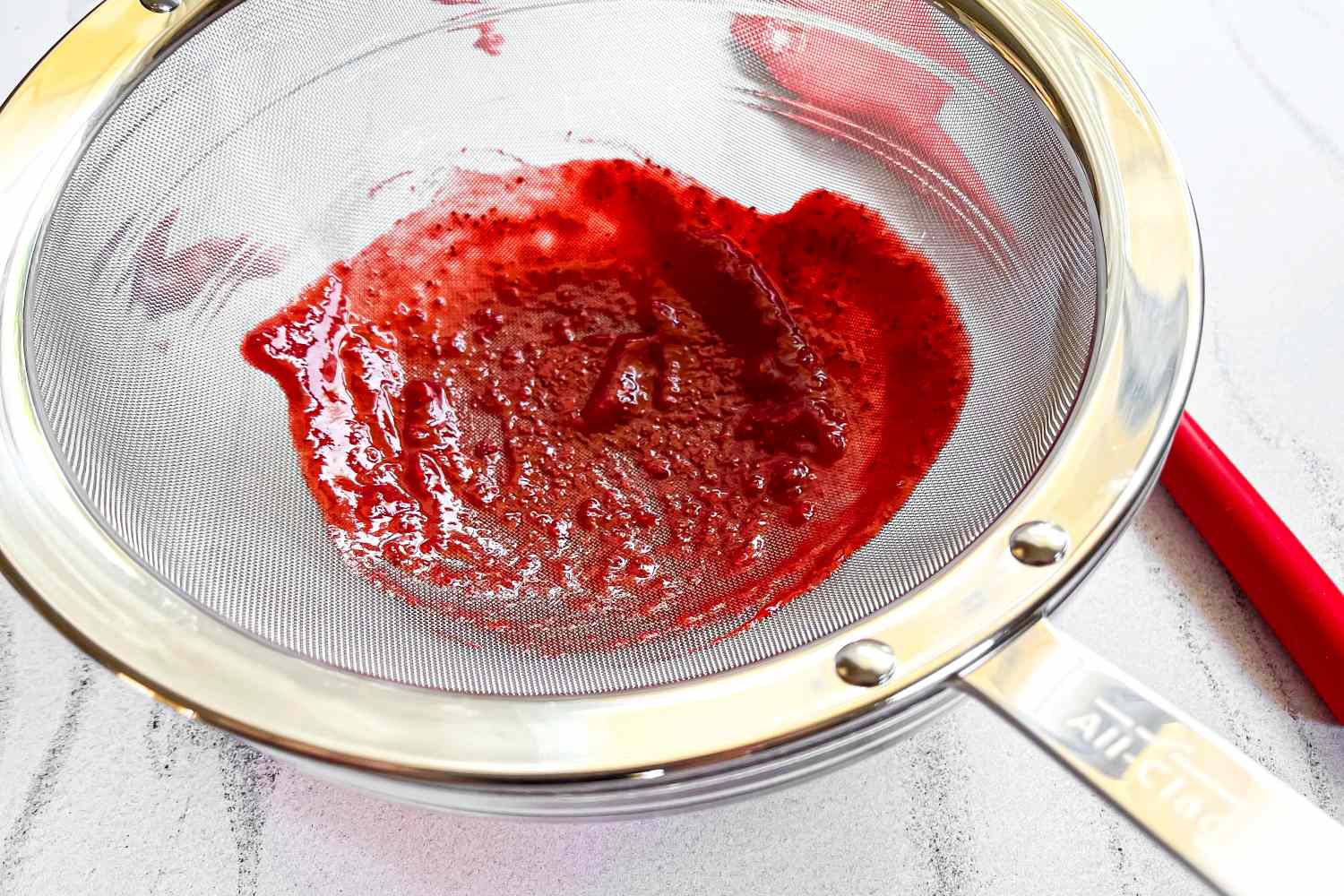 All-Clad Stainless-Steel Strainer, Set of 3 with red puree being pressed through accompanied by a kitchen utensil handle visible in the background