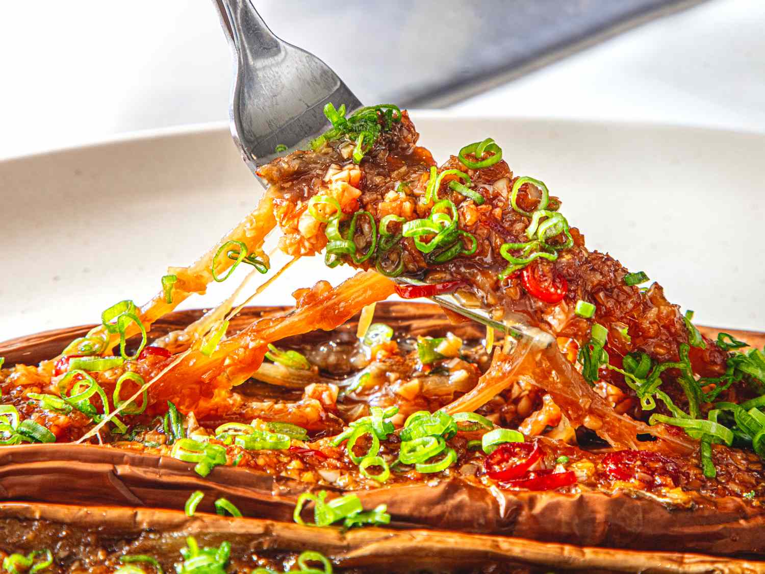 A fork lifting a piece of Chinesestyle BBQ eggplant garnished with chopped green onions on a plate
