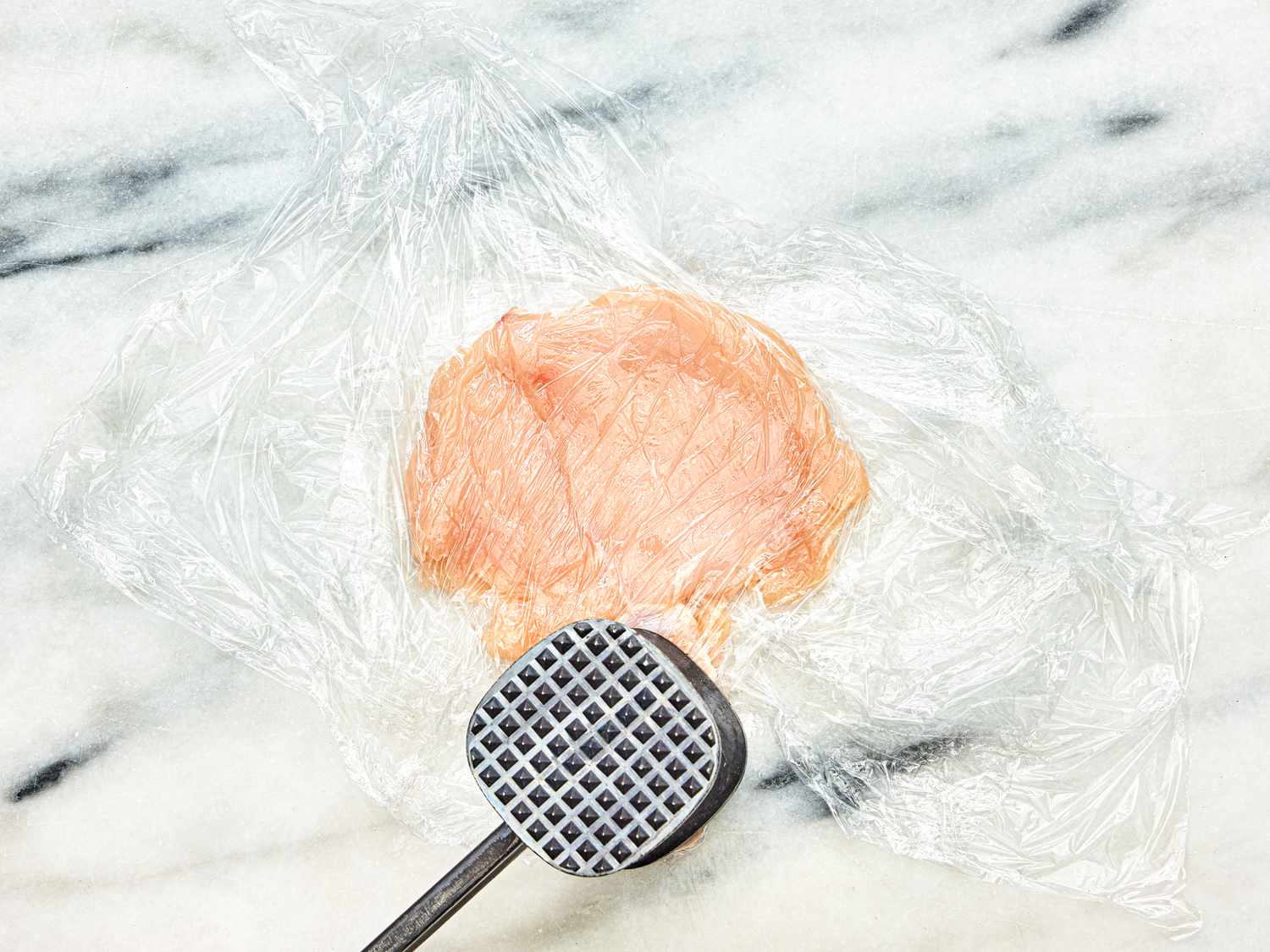 Chicken is pounded between layers of plastic wrap with a mallet on a white marble surface.