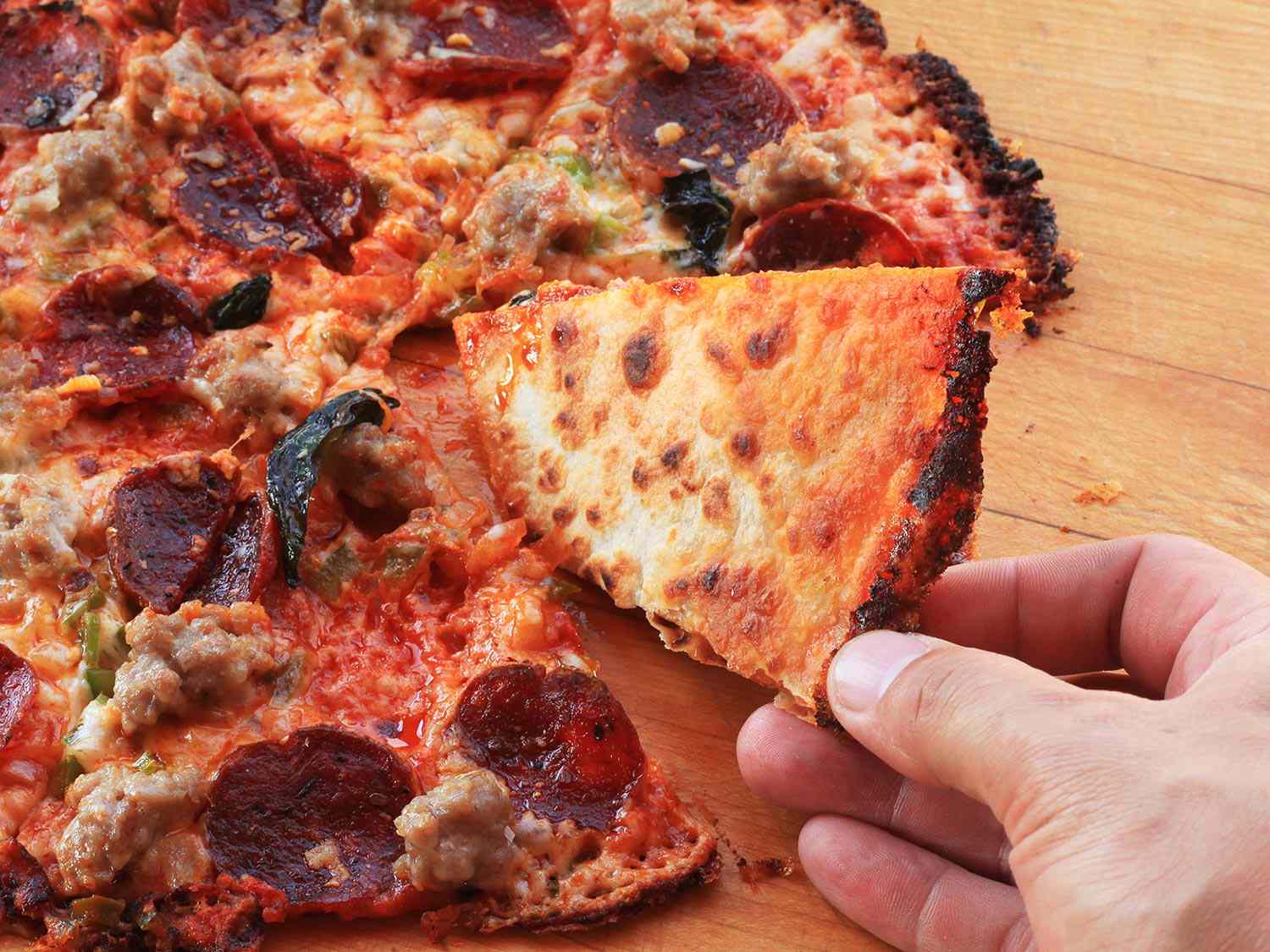Author tilting a piece of the supreme tortilla pizza over to reveal a golden, lightly charred bottom crust.