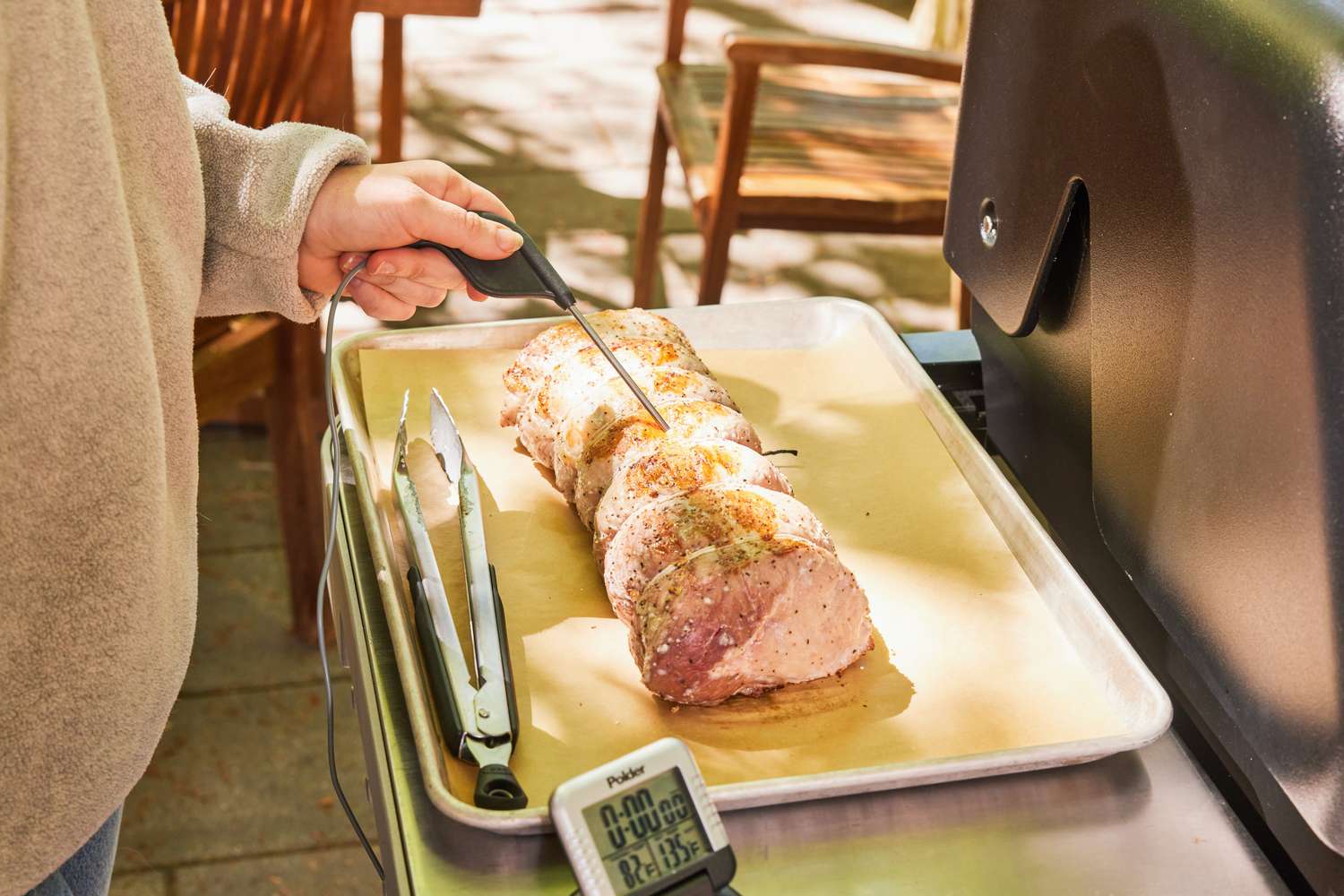 A person checking a roasted meats temperature using a digital thermometer with tongs and oven thermometer nearby on a tray