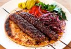 Two Adana kebabs on a plate with flatbread, sumac onions, parsley, tomatoes, and pickled peppers. 