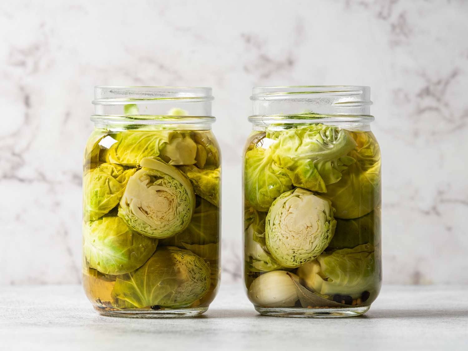Two glass jars, now filled with sliced brussels sprouts and brine. The jars are in front of a white marbled background.