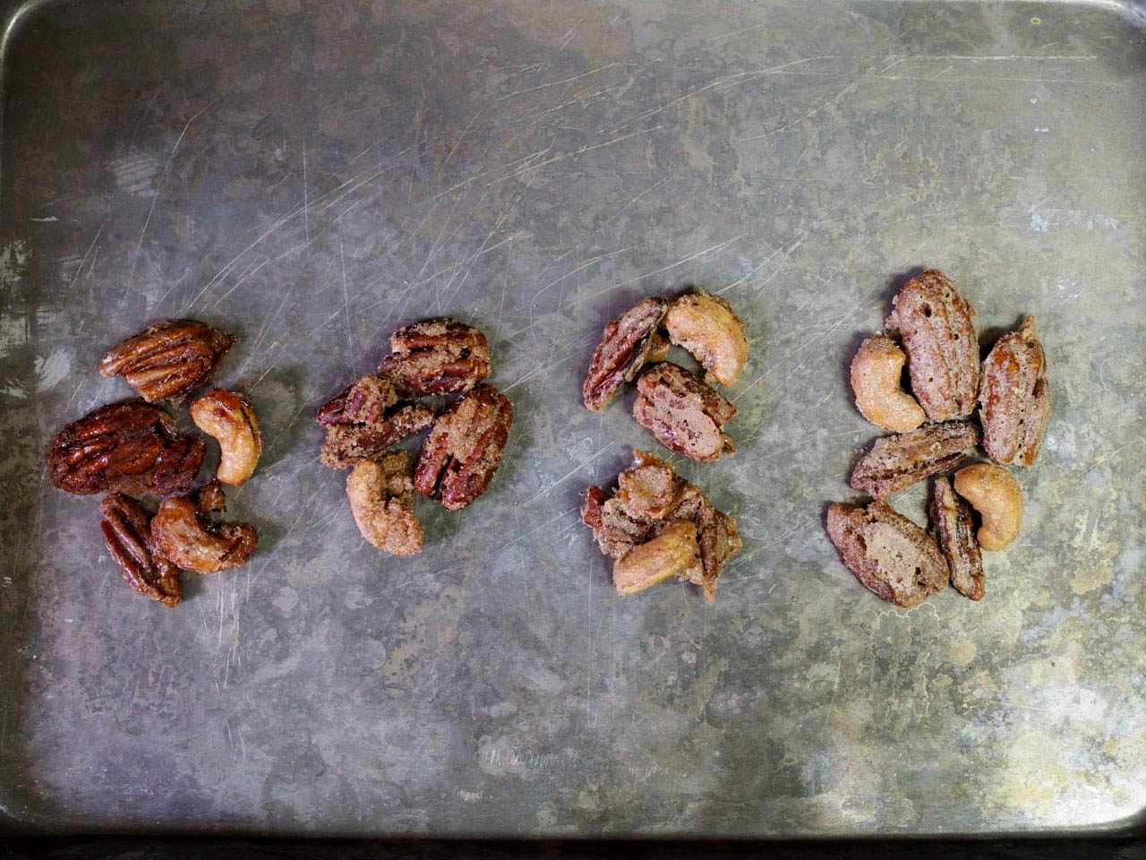 Four groups of nuts on a baking sheet with different coatings. From left to right, there are nuts have no egg, one egg per quart of nuts, two eggs per quart, and three eggs per quart.