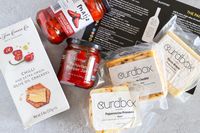 Various gourmet food items from Curdbox Cheese Subscription Box, including crackers pepper jam and cheese with labels visible from brands