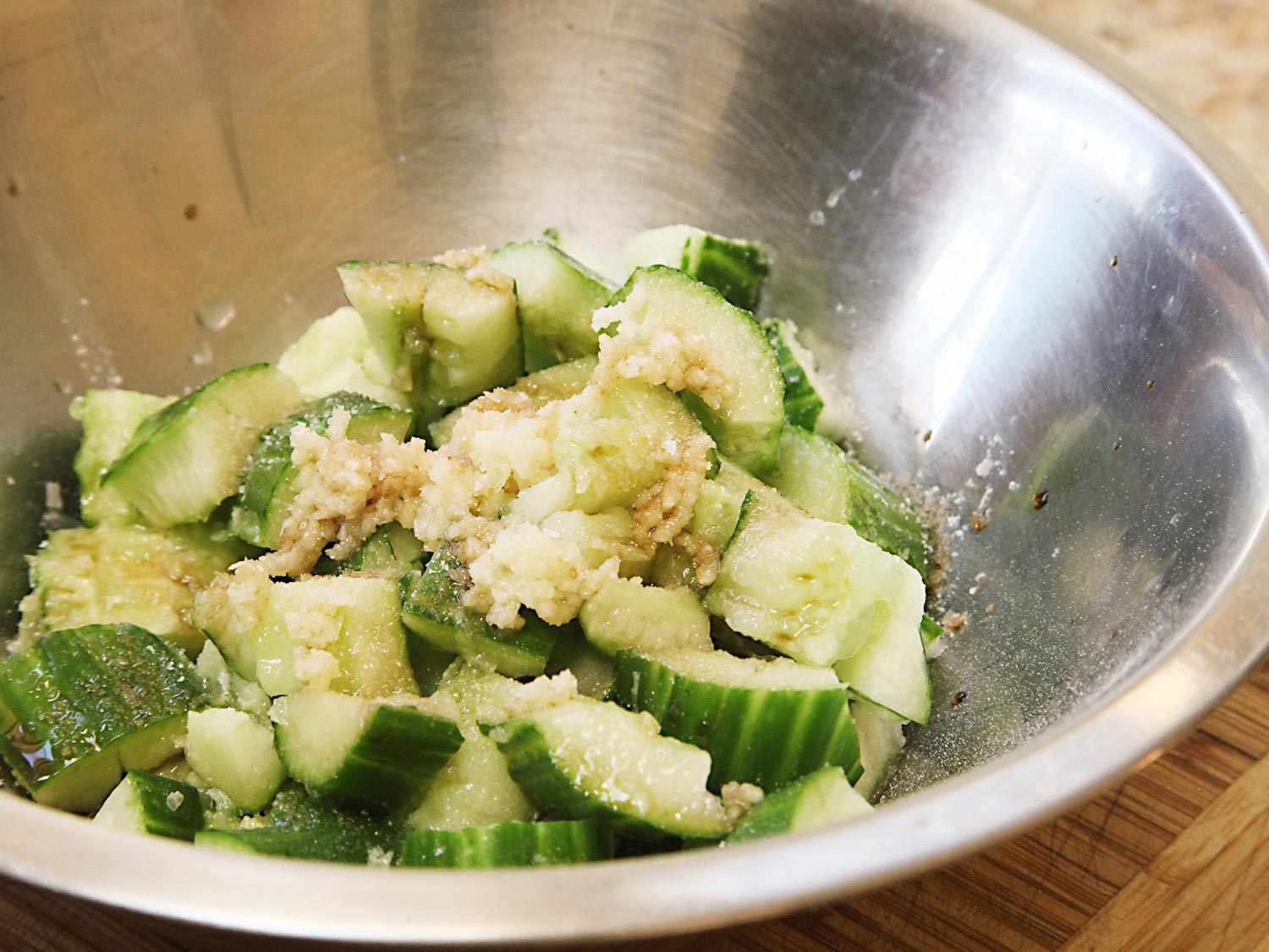 A mixing bowl containing mashed cucumber cubes, garlic, sesame oil, and vinegar. 