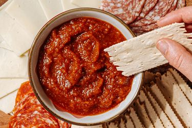 A hand holding a cracker above a bowl of red dip surrounded by slices of cheese and meats