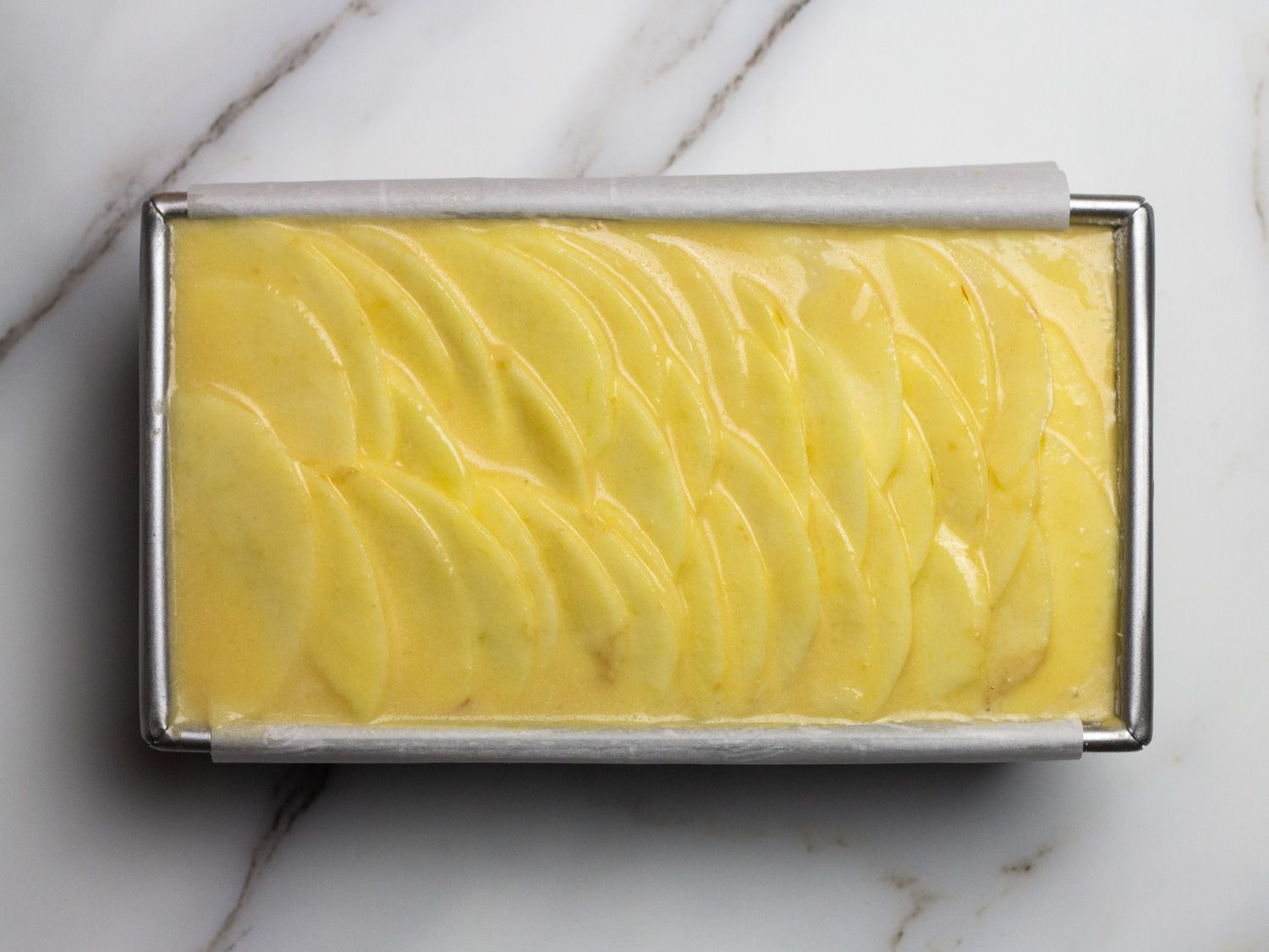 Thinly sliced apples and batter arranged in a loaf pan, ready for baking.