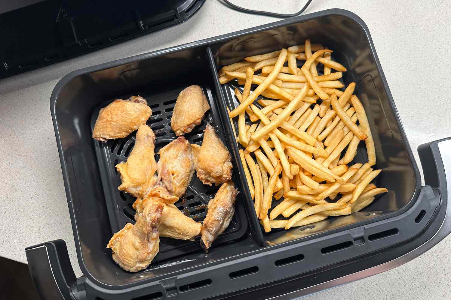 Air fryer basket with cooked chicken wings and French fries divided into two sections