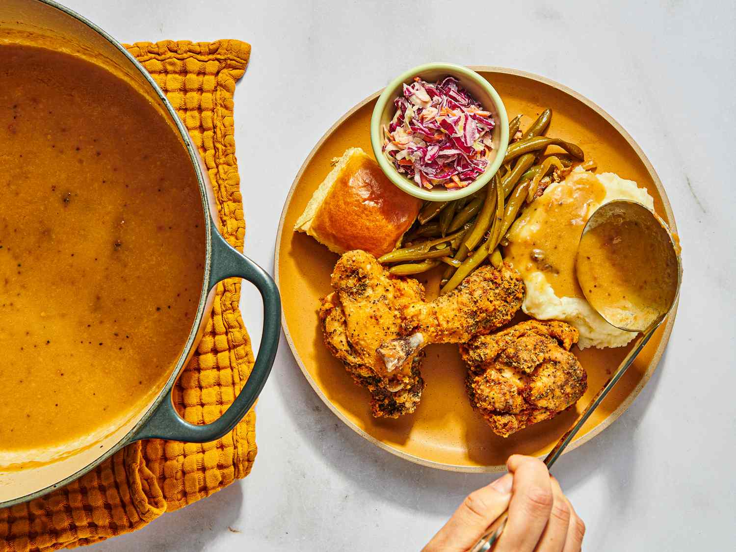 pouring gravy onto mashed potatoes on a dish with fried chicken, green beans, coleslaw