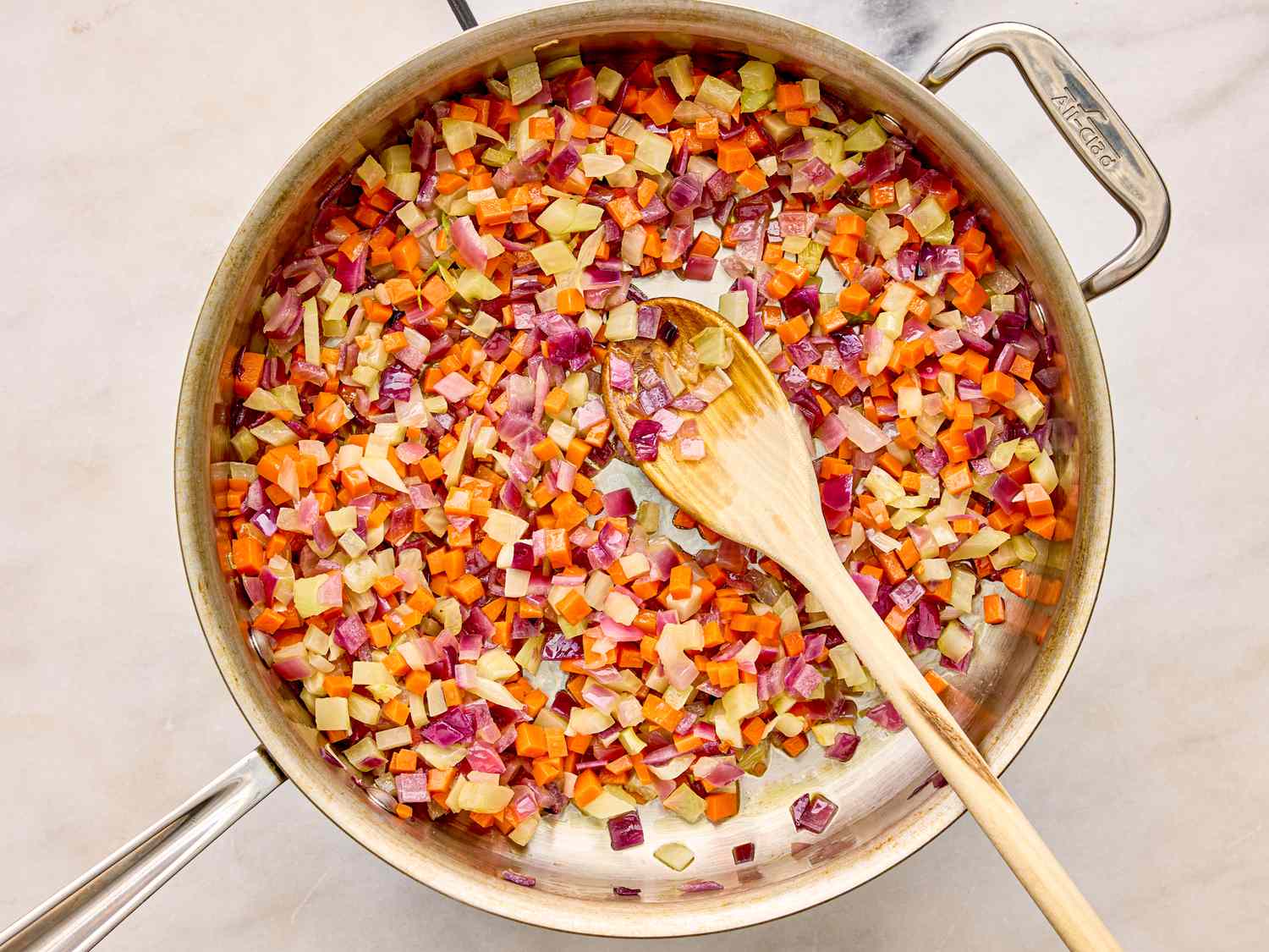 A pan with diced vegetables being sauted a wooden spoon resting in the pan