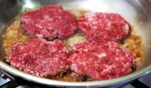 Four beef patties cooking on top of diced onion in a skillet.
