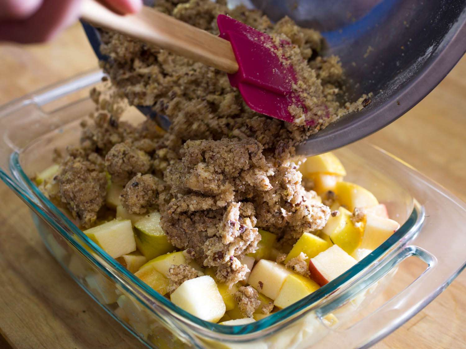 Using a flexible spatula to scrape crumb topping over chopped apples in a glass baking dish.