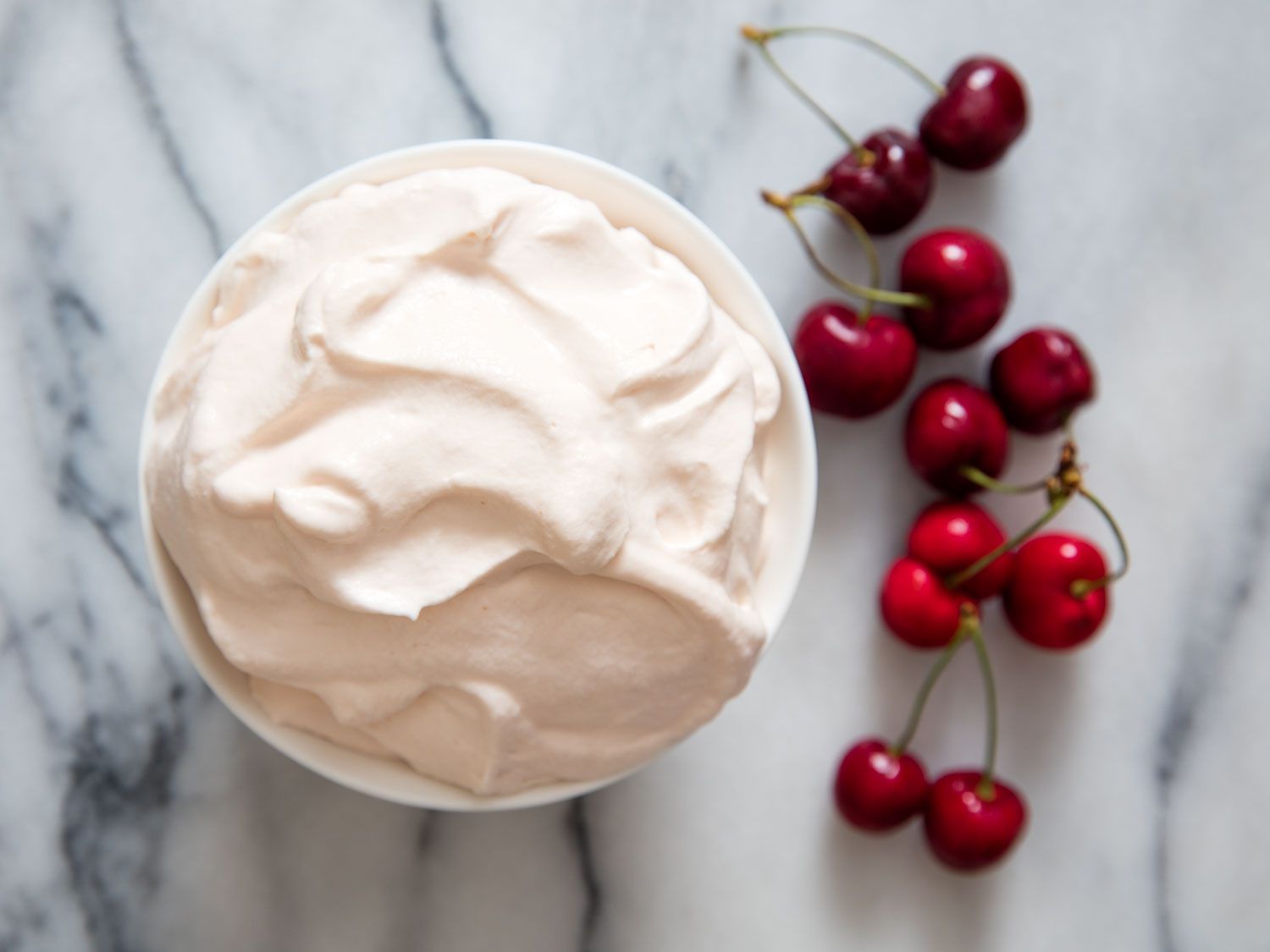 Overhead shot of cherry pit whipped cream alongside fresh cherries on marble.