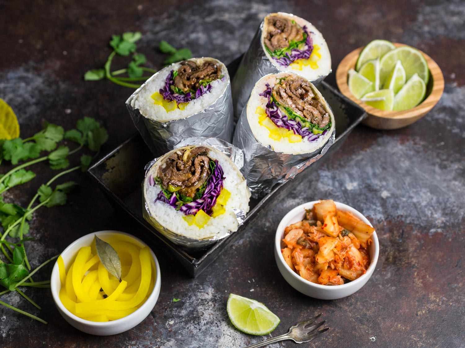 Korean bulgogi burritos displayed with bowls of lime wedges, pickled radish, and kimchi