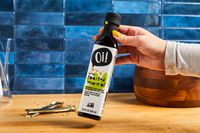 Hand holding a bottle labeled 'Oi! toasted sesame oil' on a kitchen counter with a wooden bowl and measuring spoons