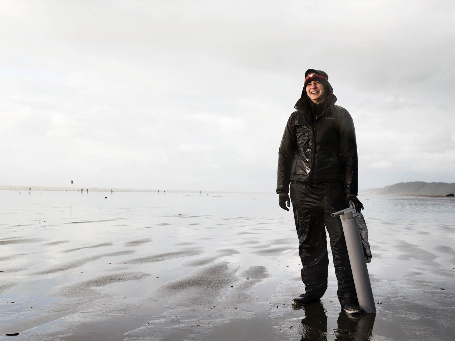 A woman dressed in rain gear standing on the beach, holding a shellfish gun.
