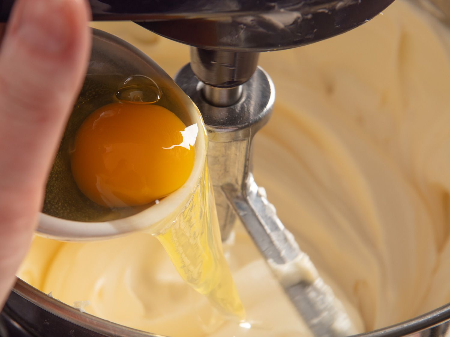An egg being poured into a cream cheese mixture that is resting in a stand mixer.