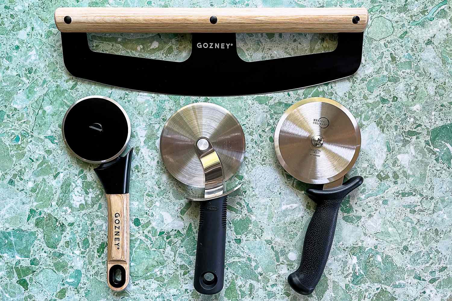 Four pizza cutters on a green surface including a large rocking blade and three rollingstyle cutters