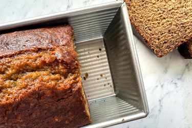 Loaf of banana bread in a metal baking pan on a marble countertop with slices of bread visible nearby
