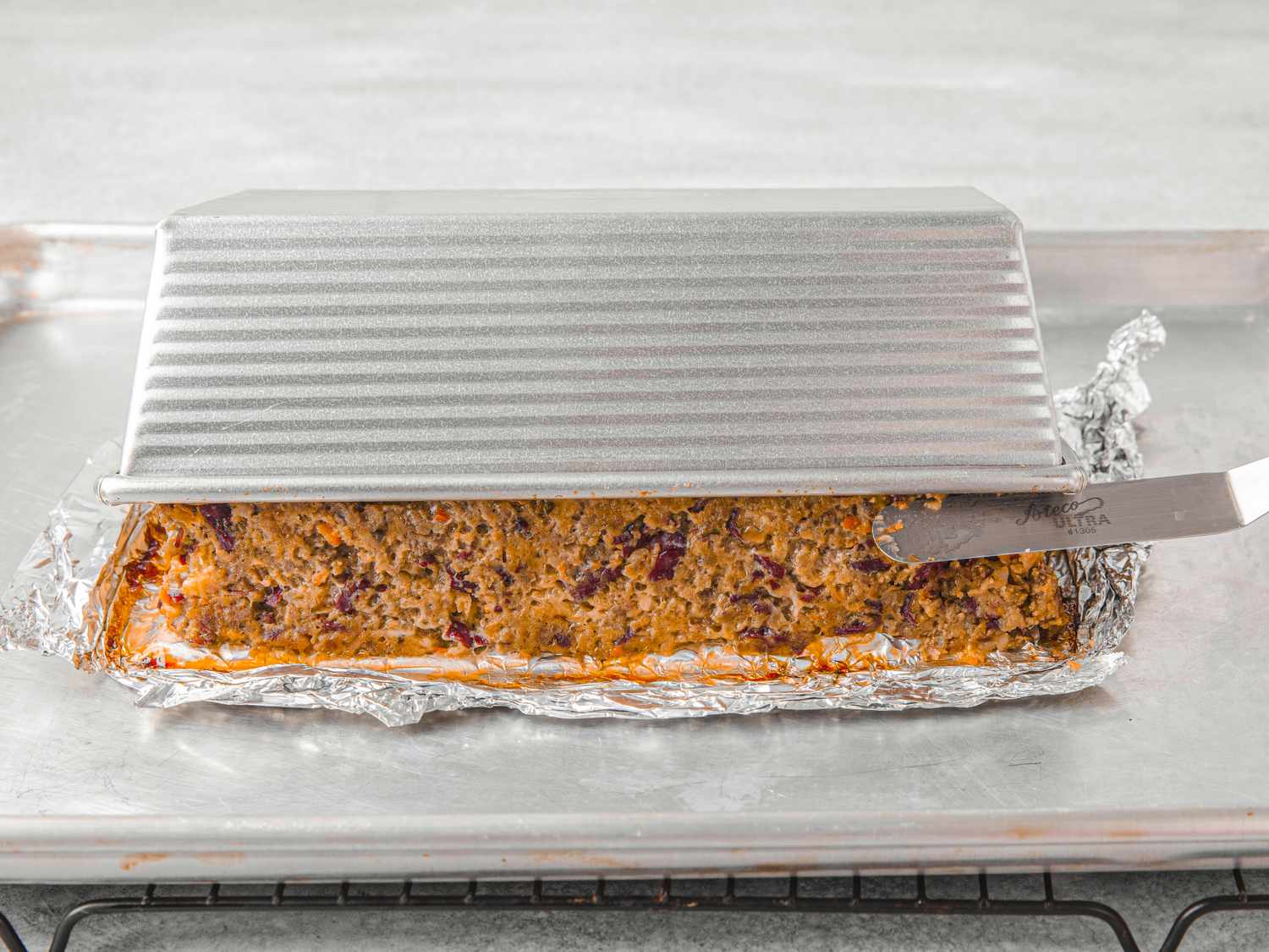 Meatloaf partially removed from a loaf pan onto a baking tray surrounded by aluminum foil cover
