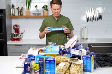 Sasha Marx from Serious Eats standing in a kitchen with a pile of different packages of pastas on the counter in front of him.
