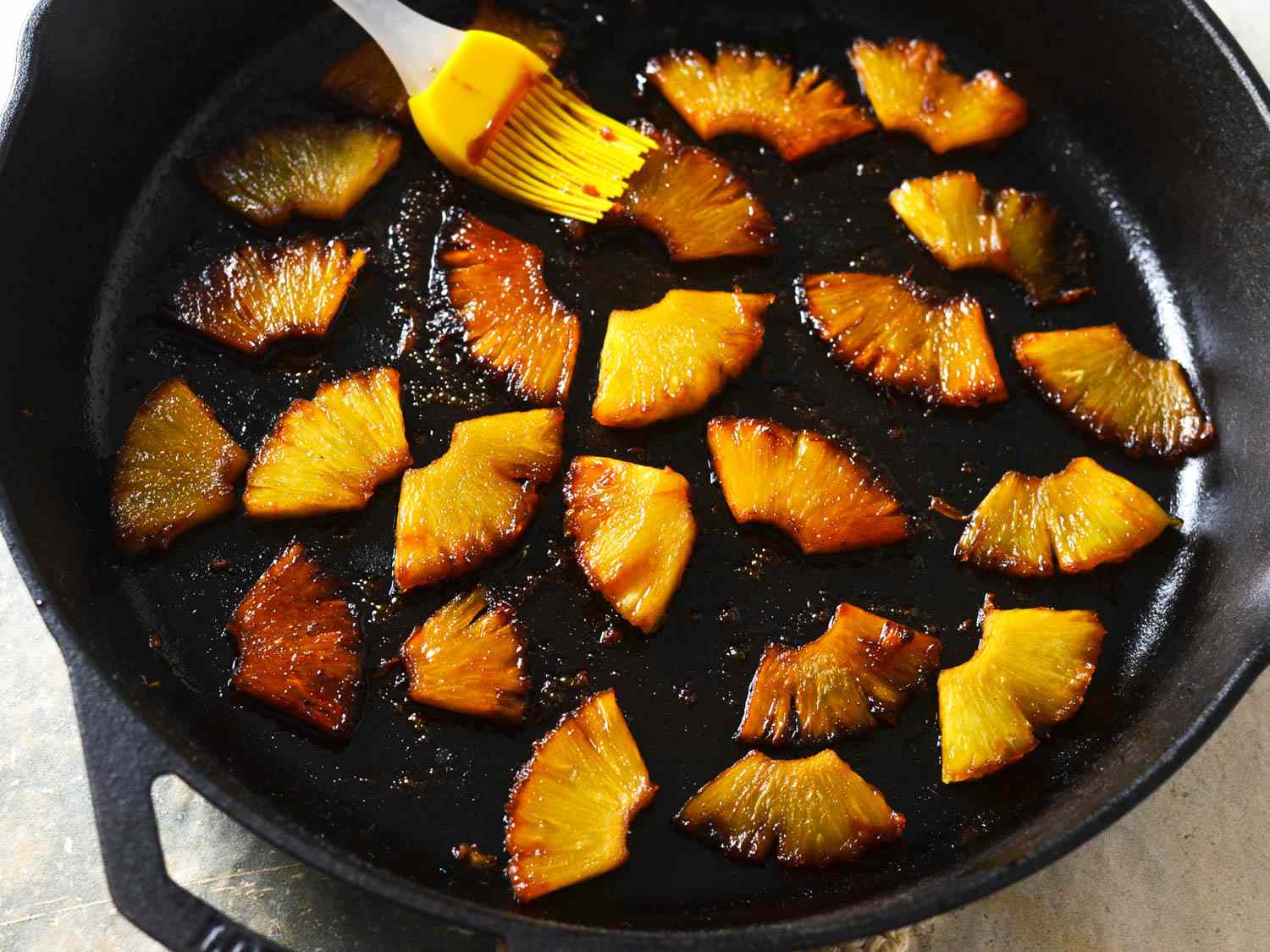 Teriyaki glaze being applied with a silicone brush on small slices of caramelized pineapple in cast iron skillet 