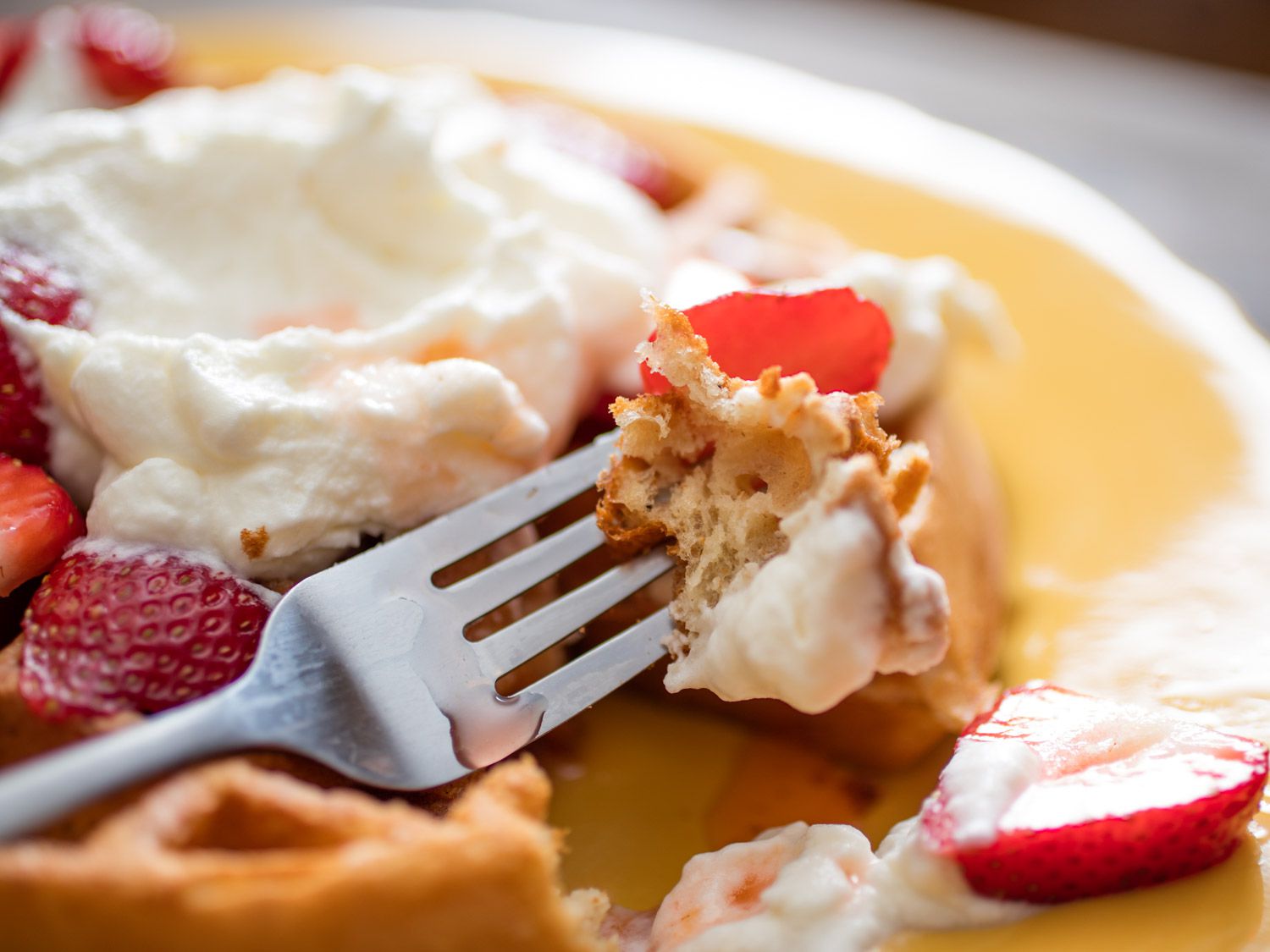 Close-up of a bite of the waffle skewered on a fork, shellacked in lemon Chantilly with a piece of strawberry balanced on top.