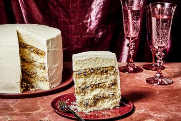 Alabama lane cake, with a slice placed on a red plate, on a red marble tabletop. 2 Magenta glasses of bubbly water, and a magenta fabric draped in the background. 