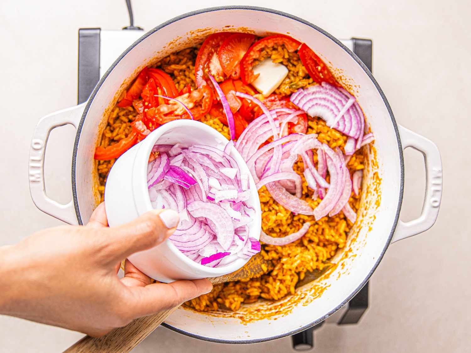 A hand adding sliced red onions into a pot with cooked jollof rice and vegetables