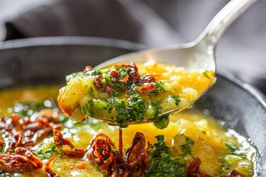 Closeup of a spoon lifting a portion of a dish with cooked rice garnished with herbs and fried shallots from a bowl