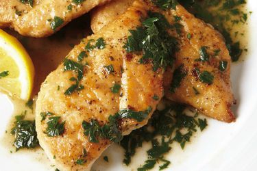 Closeup of chicken cutlets in a pan sauce with fresh herbs and a lemon wedge.