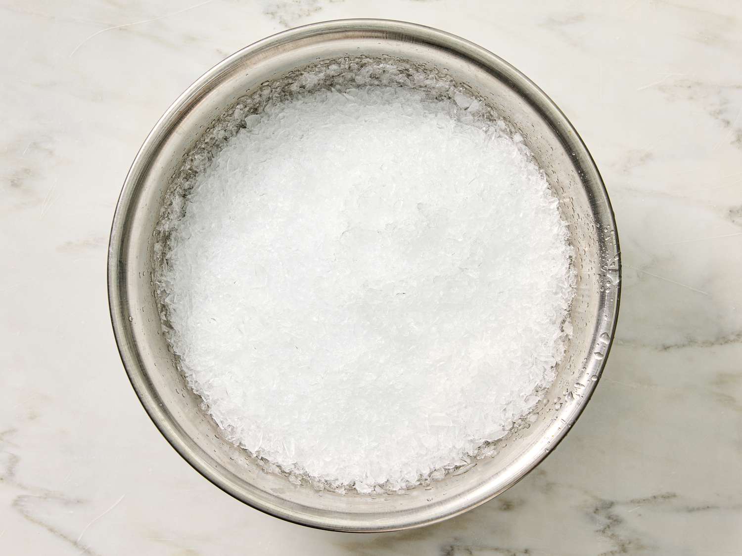 A metal bowl filled with granular white snowlike substance placed on a marble surface