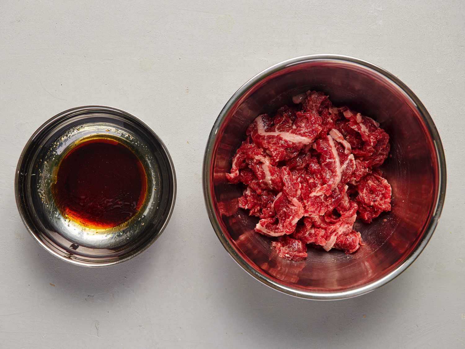 Beef, cornstarch, black pepper, Â¼ teaspoon of sugar, 1 teaspoon of Shaoxing wine, 1 teaspoon of fish sauce, and 1 teaspoon of sesame oil combined in a bowl, next to a separate bowl containing combined remaining sugar, soy sauce, fish sauce, Shaoxing wine, and sesame oil