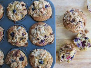 An overhead view of six berry muffins with a streusel topping in a pan on a wooden table, with one whole muffin and one split in half next to it.