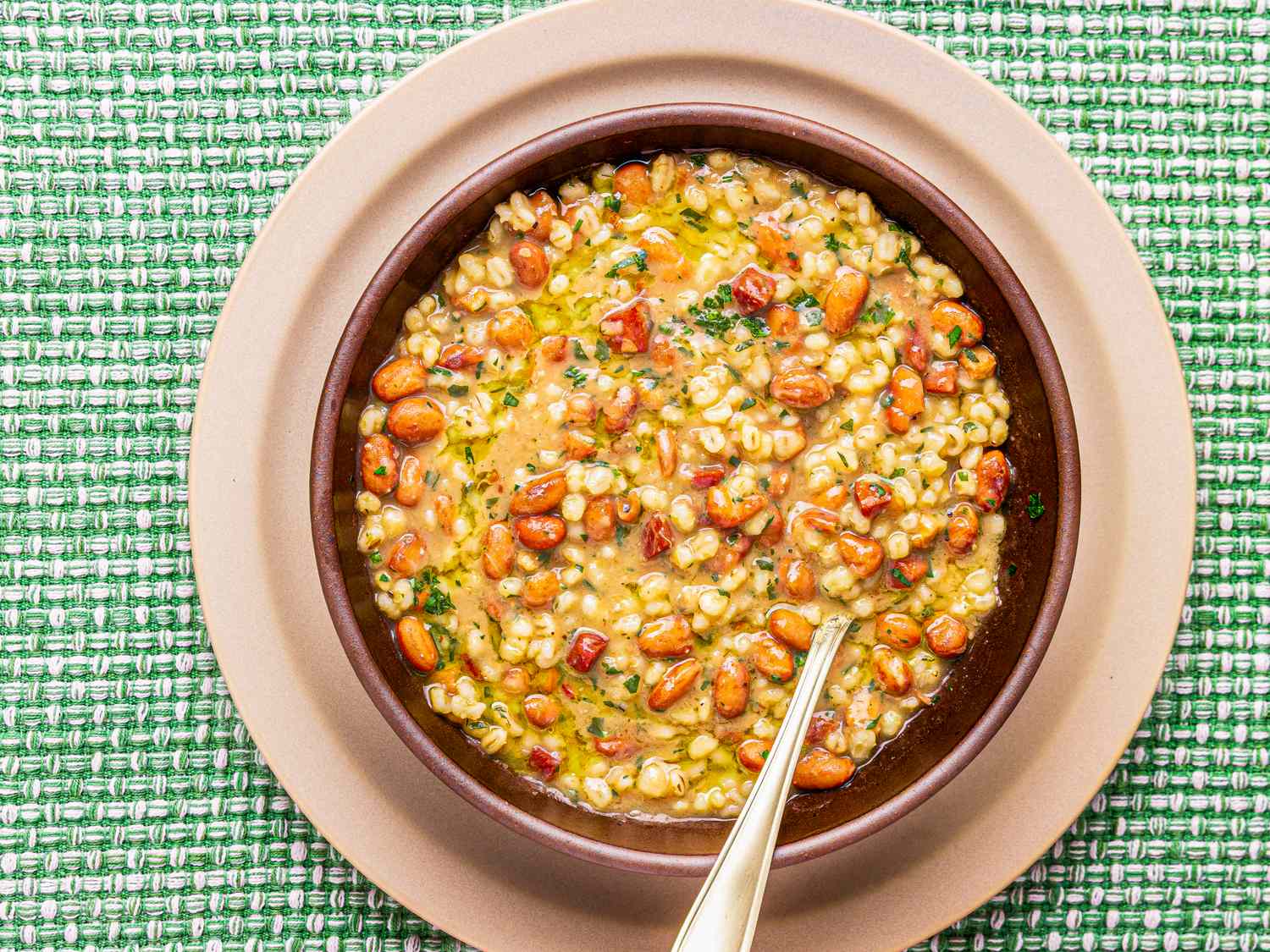 A bowl of barley and cranberry bean soup with a spoon on a green patterned background