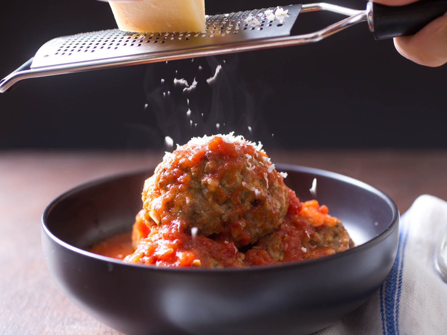 Grating hard cheese with a microplane over a dish of meatballs with tomato sauce. 